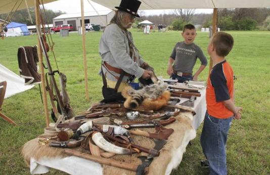 A man in frontier garb shows a table full of pelts to two boys. 