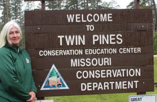 Reva Dow stands next to the entrance sign to Twin Pines Conservation Center
