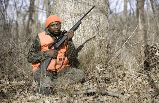 A deer hunter sits against a tree with his rifle.