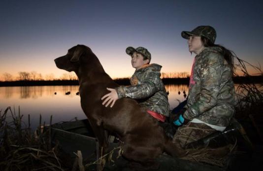 A brother and sister with their dog on a duck hunting trip.